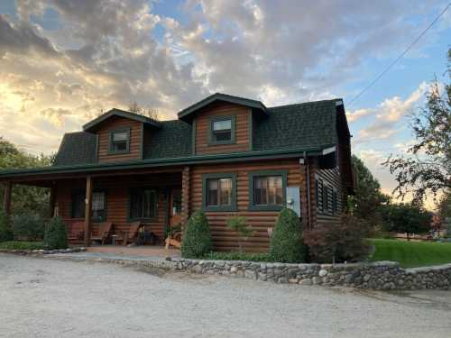 A charming log cabin with a green roof, surrounded by trees and a gravel path, under a cloudy sky at sunset.