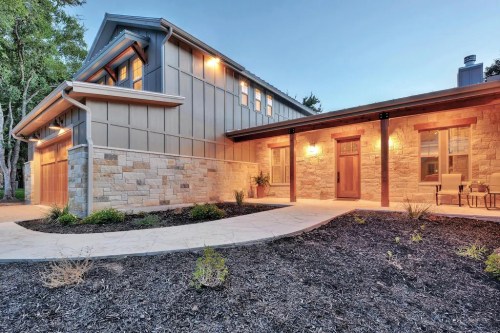 A modern home exterior with stone and wood accents, illuminated by warm lights, surrounded by landscaped greenery.