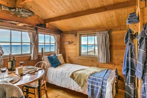 Cozy cabin interior with a bed, dining table, and large windows overlooking a serene lake view.