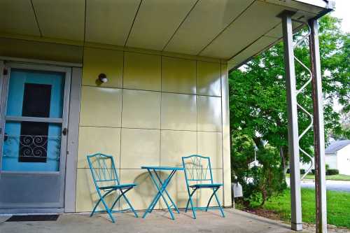 A small patio with a blue table and two chairs, next to a light yellow wall and greenery in the background.