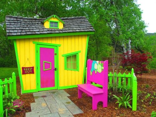 Colorful, whimsical garden shed with a bright yellow exterior, pink accents, and a matching bench in a lush green setting.