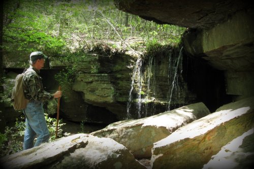 A person in camouflage stands near a rocky area with a small waterfall surrounded by lush greenery.