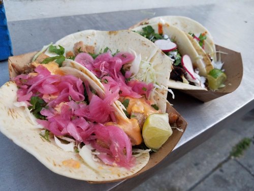 Two taco platters with tortillas filled with meat, pink pickled onions, lettuce, radishes, and lime wedges on a metal surface.