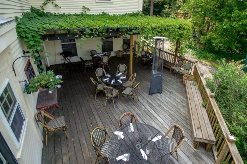A wooden patio with tables and chairs, shaded by greenery, surrounded by trees and a peaceful outdoor setting.