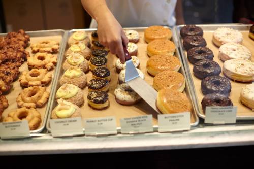 A hand lifts a donut from a display of various donuts, including glazed, chocolate, and filled varieties.