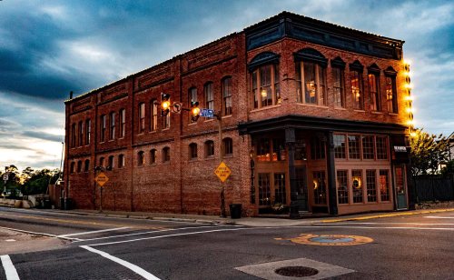 Historic brick building at dusk, featuring large windows and warm lights, located at a street intersection.