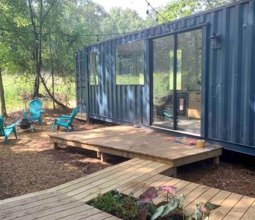 A blue shipping container home with a wooden deck, surrounded by greenery and outdoor seating.