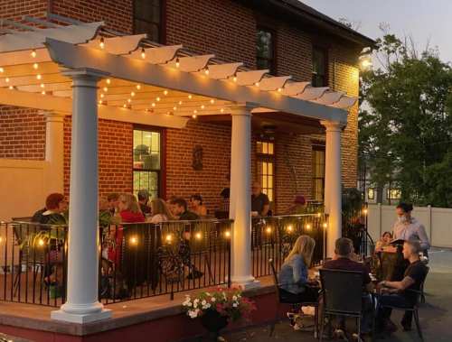 A cozy outdoor dining area with string lights, featuring groups of people enjoying a meal on a warm evening.
