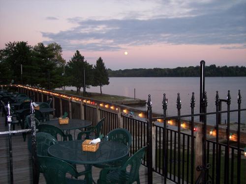 A serene lakeside view at dusk, featuring a deck with tables, string lights, and a rising moon over the water.