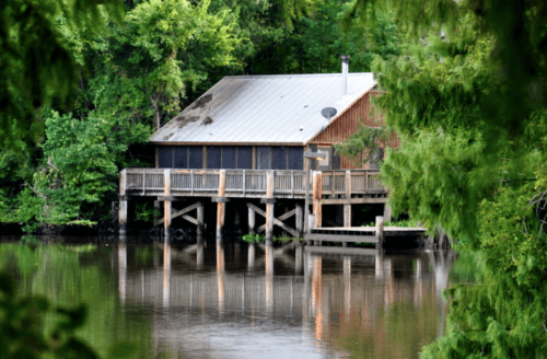 A rustic cabin on stilts by a calm river, surrounded by lush greenery and reflecting in the water.
