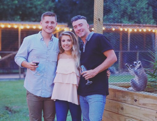 A group of three friends poses for a photo outdoors, with a raccoon visible in the background near a wooden fence.