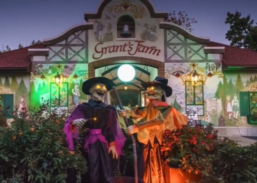 Two spooky skeletons in costumes stand outside Grant's Farm, illuminated by colorful lights against a festive backdrop.