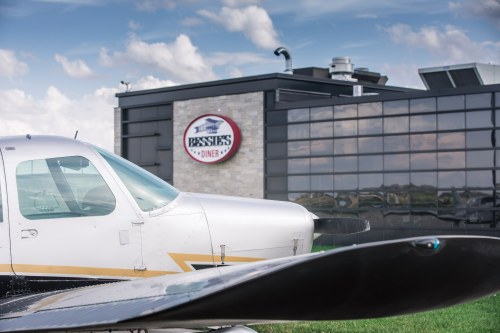 A small airplane in front of a modern diner with a sign reading "Bessie's Diner" against a cloudy sky.