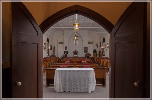 View through arched doors into a church interior with wooden pews and a table at the front, decorated with a white cloth.