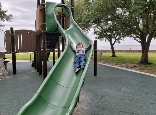 A child joyfully sliding down a green playground slide, with trees and a grassy area in the background.