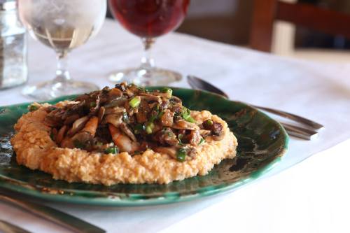 A green plate with creamy polenta topped with sautéed mushrooms and herbs, set on a table with drinks in the background.
