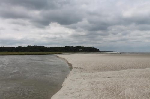 A sandy beach curves along a riverbank under a cloudy sky, with trees in the background and a few distant figures.