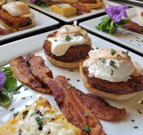 Plates of eggs Benedict with poached eggs, bacon, and garnishes, served alongside a cheesy potato dish.