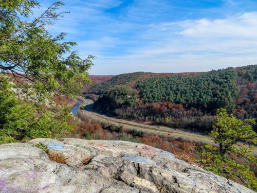 A scenic view of a river winding through green hills under a blue sky, with rocky foreground and autumn foliage.