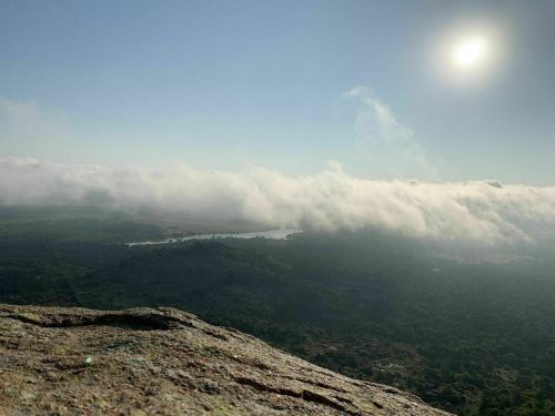A panoramic view from a rocky peak, with clouds rolling over a valley and a bright sun in the sky.