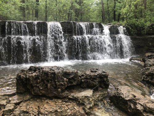 A serene waterfall cascades over rocky ledges, surrounded by lush green trees in a tranquil forest setting.