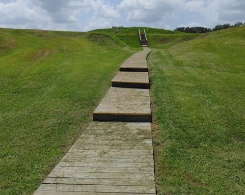 A wooden pathway leads up a grassy hill, with steps at the top under a partly cloudy sky.