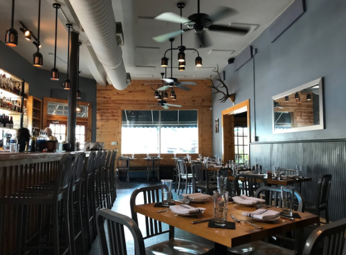 A modern restaurant interior with wooden accents, bar seating, and tables set for dining, featuring ceiling fans and large windows.