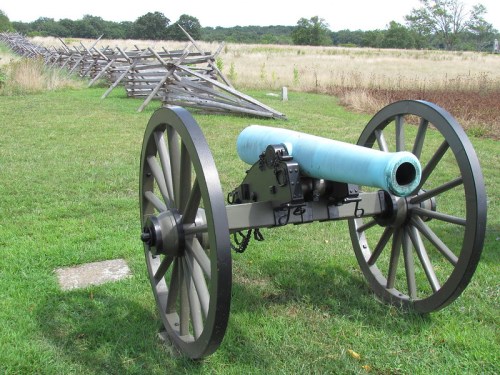 A historic cannon on wheels positioned on grassy terrain, with a wooden fence line in the background.