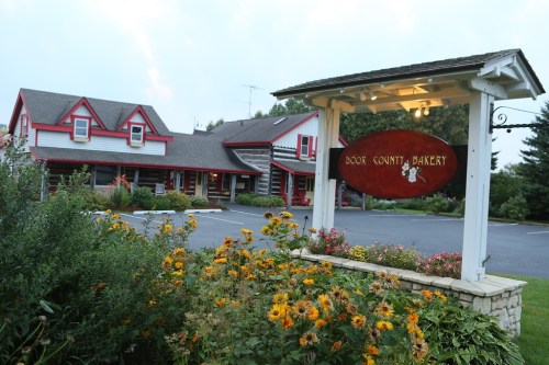 A charming bakery with a red sign, surrounded by colorful flowers and a scenic building in the background.