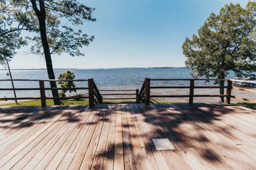 A wooden deck overlooking a calm lake, surrounded by trees under a clear blue sky.