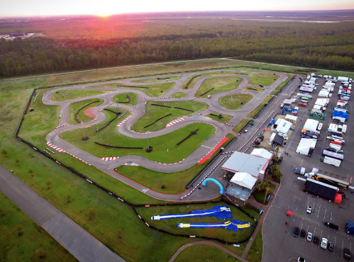 Aerial view of a racetrack surrounded by trees, with a paddock area filled with tents and vehicles at sunset.