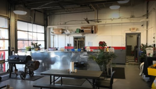 Interior of a spacious room with a metal counter, plants, a grill, and a dining table, featuring large windows and bright lighting.