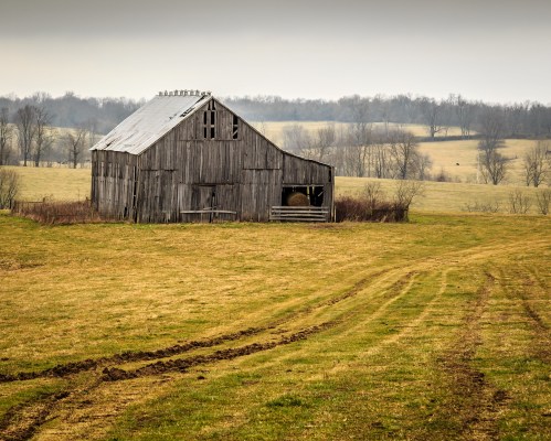 A weathered barn stands in a grassy field, surrounded by trees and a cloudy sky, with tire tracks leading toward it.