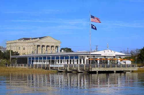 A waterfront restaurant with a deck, flags flying, and a historic building in the background against a clear blue sky.