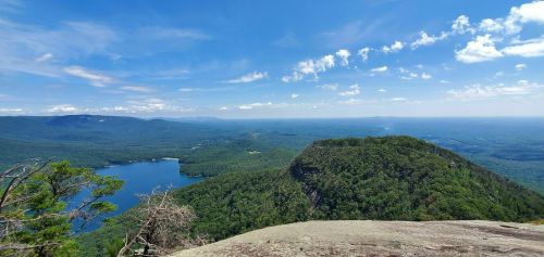A panoramic view of lush green mountains and a blue lake under a bright sky with scattered clouds.