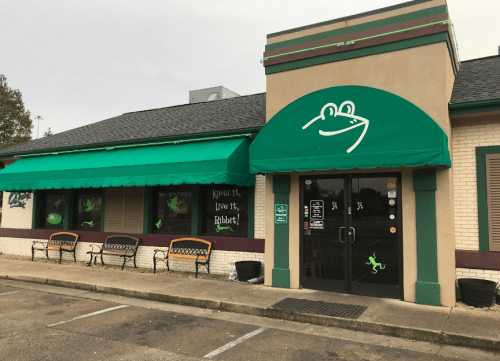 A restaurant exterior with a green awning, frog logo, and benches outside, featuring a playful and inviting design.