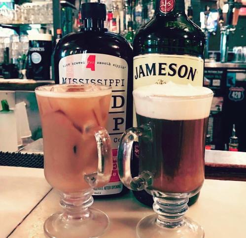 Two glasses of drinks on a bar counter, one with iced coffee and the other with a creamy beverage, alongside whiskey bottles.