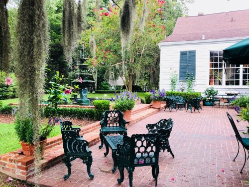 A serene garden with a fountain, surrounded by lush greenery and decorative seating on a brick patio.
