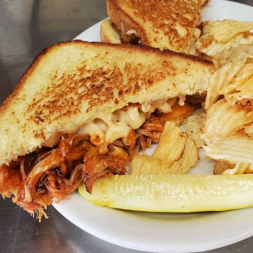 A plate featuring a grilled cheese sandwich with pulled chicken and macaroni, served with potato chips and a pickle.