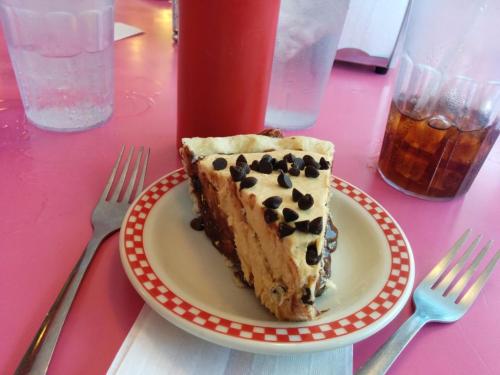 A slice of dessert with chocolate chips on a plate, accompanied by forks and drinks on a pink table.