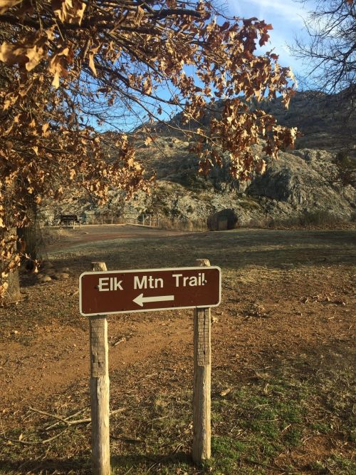 A brown sign pointing left, indicating the direction of the Elk Mountain Trail, with a rocky landscape in the background.
