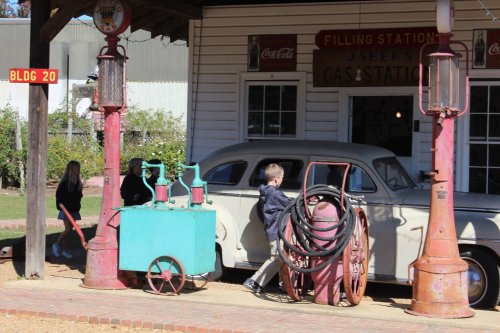A vintage gas station with a classic car, a child interacting with old gas pumps, and a retro filling station sign.