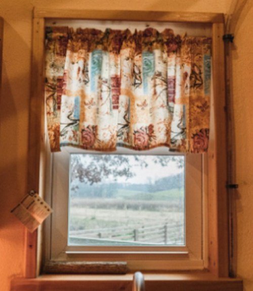 A window with a floral curtain, revealing a view of a foggy landscape and distant fields.
