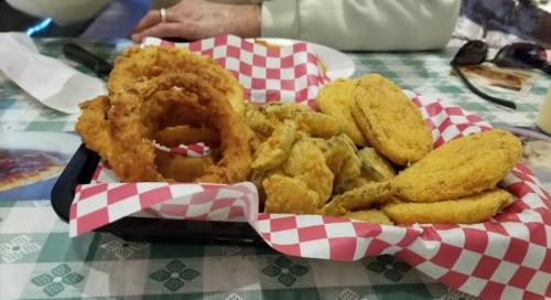 A basket of fried food including onion rings, fried pickles, and corn fritters on a checkered paper liner.