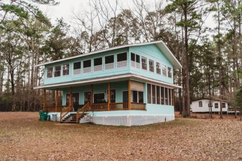 A two-story light blue house with a porch, surrounded by trees and fallen leaves in a wooded area.