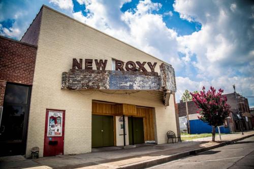 Exterior of the New Roxy theater, featuring a weathered marquee and a bright blue sky with fluffy clouds.