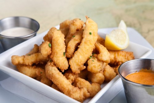 A plate of crispy fried fish strips served with lemon wedges and two dipping sauces.