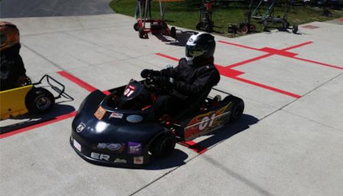 A go-kart racer in a black helmet and suit sits in a go-kart on a paved track with red markings.