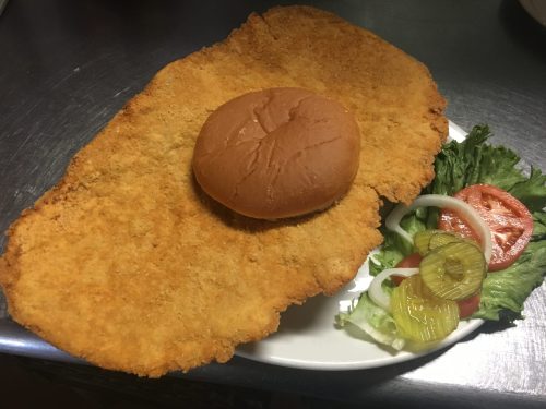 A large breaded cutlet on a plate with a bun and a side of lettuce, tomato, and pickles.