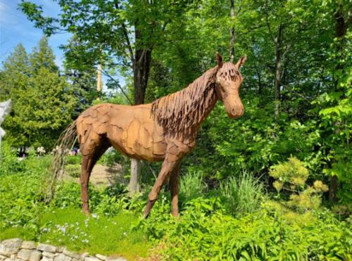A life-sized sculpture of a horse made from wood, surrounded by greenery and flowers in a sunny outdoor setting.